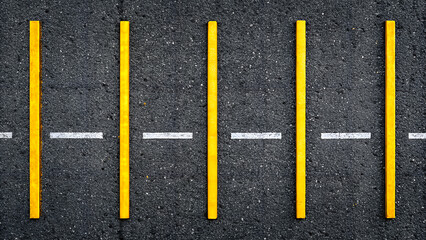 A top down view of a paved asphalt surface with a white dashed line and six evenly spaced bright yellow vertical poles marking a boundary or pathway