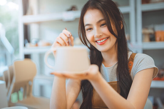 Asian woman painting ceramic art and pottery designs in her studio – happy artisan potter sitting with handmade tools, enjoying sculpting process and creative small business lifestyle