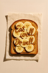 Healthy Toast with Banana and Peanut Butter, Inspirational Quote “Eat Well. Do Well.” Written in Syrup – Wholesome Food Typography Concept, Editorial Top View on Beige Background