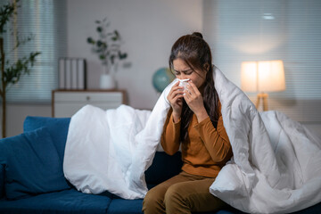 Sick asian woman blowing nose while sitting on sofa covered in blanket at night