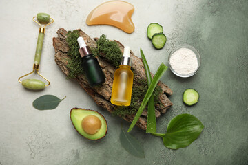 Composition with bottles of cosmetic products, facial massage tools, tree bark and ingredients on green grunge background