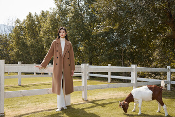 Young woman in a coat standing in a grassy area by a fence.