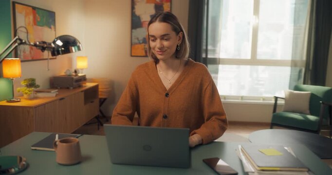 Beautiful Caucasian Specialist Working on Laptop Computer in a Stylish Living Room at Home. Sitting Behind a Desk. Freelance Female Browsing Internet, Using Social Networks, Feeling Happy. Arc Shot