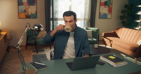 Middle Eastern Young Man Working Remotely from Home, Sitting in a Living Room Behind a Desk and Using a Computer. Male is Drinking from a Cup while Browsing Reports a Laptop. Creative Arc Footage - Powered by Adobe