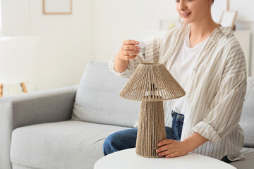 Young woman screwing light bulb into lamp on table at home