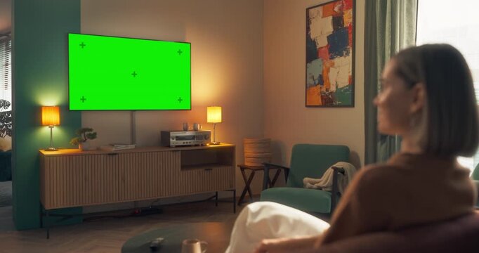 Zoom Out with Speed Ramp of a Female Sitting on a Sofa and Watching TV. Television Set at Home with a Green Screen Chroma Key Template with Placeholders. TV Hanging on the Wall in the Apartment