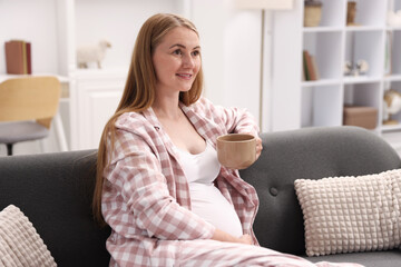 Beautiful pregnant woman with cup of drink on sofa at home