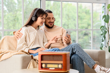 Young couple in headphones with mobile phone listening to radio on sofa at home