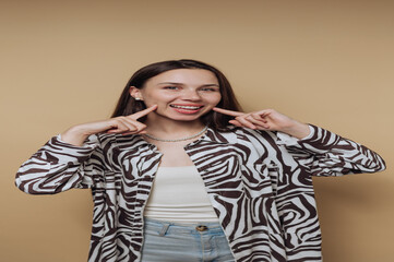 portrait of young woman poses joyfully, pointing at her cheek while smiling broadly on calm yellow...