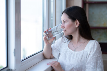 Peaceful woman enjoying a glass of water while looking out the window. Hydration and mindfulness at...