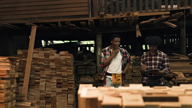 Factory Worker Inspecting Timber And Wood Furniture As Two African American Male Woodworkers Discuss Stock Checking And Quality Inspection In Industrial Workshop