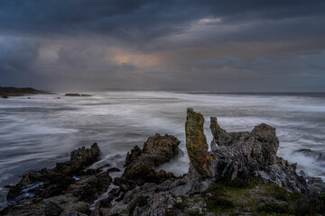 Powerful waves crash onto rugged rocks under dramatic storm clouds along the Hermanus coastline, Whale Coast, Overberg, Western Cape, South Africa — capturing the wild beauty of the seascape.