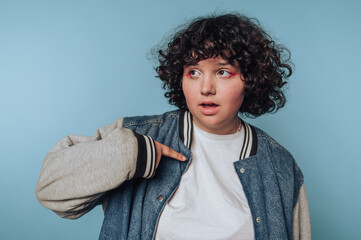 portrait of young woman points to their jacket, reflecting excitement and wonder on blue background