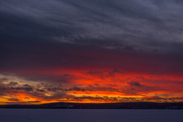 Fototapeta premium A dramatic winter sunset over a frozen lake. Dazzling shades of red, orange and purple break through heavy grey-blue clouds, painting the sky above the horizon in fiery colors. 