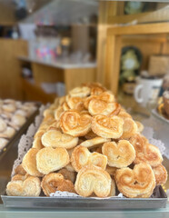 A detailed close-up of bunch of heart-shaped Palmiers (also known as elephant ears or French hearts). The background is blurred, suggesting a bakery display, a kitchen counter, or a pastry shop,