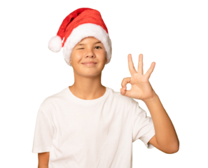 Portrait of a smiling boy in a red santa Claus hat and white t-shirt with okay sign gesture on transparent background.