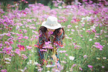 A woman in a hat leans down to smell the sweet fragrance of cosmos flowers in a vibrant field. A...