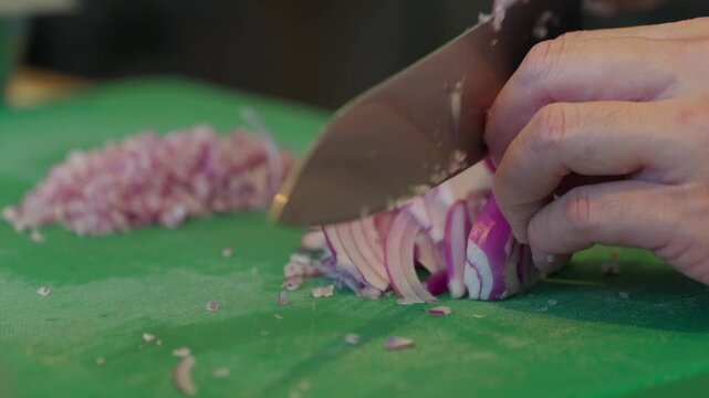 Close up, A chef slices a red Spanish onion with a very sharp black handled chef knife sliding it off the knife onto the green chopping board ready to finely dice.