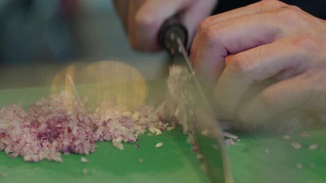 Close up, A chef finely dices thinly sliced purple onion on a green chopping board with a sharp black handled chefs knife.