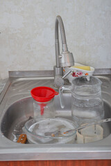 dirty dishes with soap suds and utensils in kitchen sink next to jug and funnel under metal faucet household chores, cleaning routine, domestic life, kitchen organization 