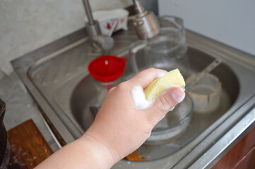 hand holds foamy sponge while washing dishes in kitchen sink with dirty bowl and utensils cleaning products, home hygiene, domestic chores, everyday life