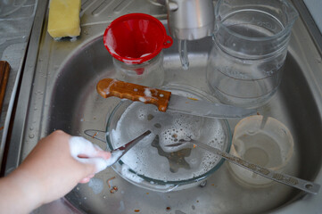 hand holds foamy sponge while washing dishes in kitchen sink with dirty bowl and utensils cleaning...