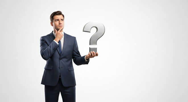 Confused businessman contemplates question mark held in hand, deep in thought. Thoughtful businessman in suit ponders problem, solution, or answer. - Powered by Adobe