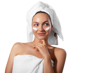Smiling young woman applying facial cream under her eyes, wrapped in a white towel with a towel...