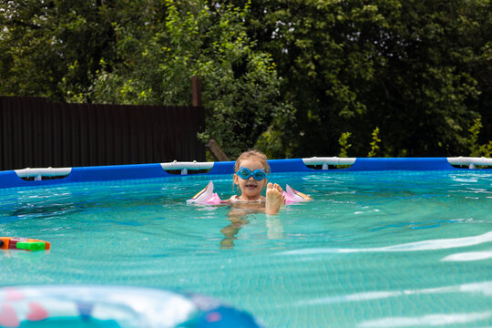 Young girl wearing goggles and armbands enjoying a sunny day while relaxing in a backyard above-ground swimming pool, surrounded by greenery and the cheerful atmosphere of summer fun.