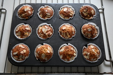A batch of freshly baked almond muffins cools in a tray lined with white paper cups. The embodiment of home baking, warm desserts, and cosy moments in the kitchen.