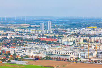 Expansive aerial view of Vienna reveals a blend of modern high-rises, residential blocks, and green landscapes. City stretches toward the horizon, framed by wind turbines and distant hills