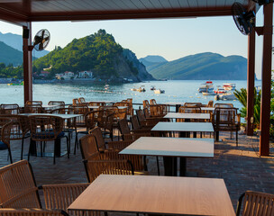 cozy outdoor cafe or restaurant located on embankment with panoramic view of Petrovac city, Montenegro, tables and chairs in front of sea and beach, beautiful nature with hills and forest