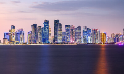 Obraz premium Sunset view of Doha skyline, Qatar, with shades of pink and purple filling the sky. Modern skyscrapers are reflected in the calm waters of the bay, as city lights begin to glow