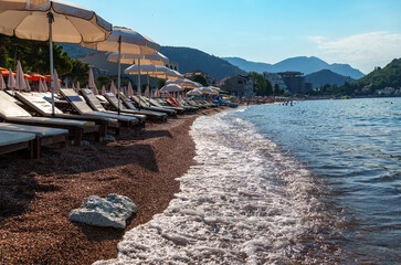 city beach with sun loungers and umbrellas with beautiful coastline and view of Adriatic sea in Petrovac, Montenegro, with high forested hills in the background