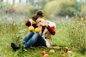 Thanksgiving Vibes. Young girl sitting on blanket in autumn meadow holding two small pumpkins, surrounded by wildflowers. warmth and whimsy of autumn harvest, childhood, and outdoor seasonal fun