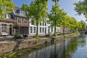 Obraz premium A serene canal scene in Amersfoort, lined with charming traditional Dutch houses and vibrant green trees, reflecting in the calm water under a clear sky.