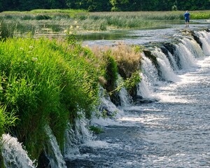 Small cascading waterfall in a lush green landscape with a person walking on top of the waterfall. The Venta River in Kuldiga in Latvia.