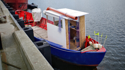 Small fishing boat docked at a pier, painted in blue and red, with a simple wooden cabin and fishing gear including floats and neon flags.