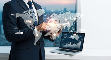  businessman using a tablet with a digital world map and financial data overlay, laptop showing charts in the background