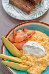 A delicious breakfast bowl featuring creamy scrambled eggs, smoked salmon, avocado slices, and a dollop of herbed yogurt served with whole grain toast.