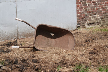 Dilapidated wheelbarrow sitting on hay-covered ground with grey panel and brick wall backgrounds...