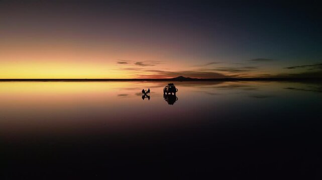 Drone footage circling 4x4 vehicle and two people on chairs at salt flats during sunset. Perfect mirror reflection on water surface. Mountains silhouetted on horizon at dusk.