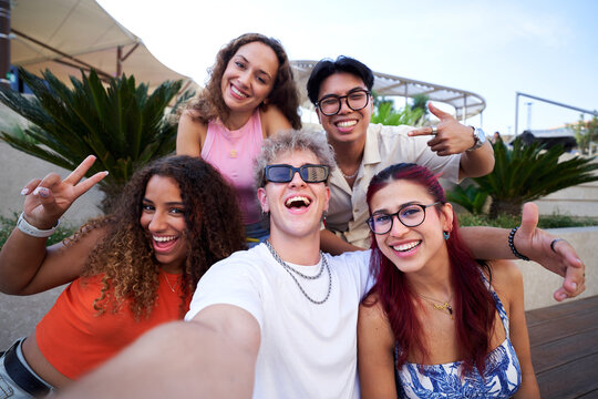 Five cheerful diverse friends taking a selfie photo while making funny faces, gathering together outdoors. Multiracial group of young people sharing memories on social media.