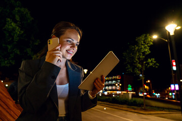 Urban young adult multitasking woman using digital tablet and talking on mobile phone at city street at night
