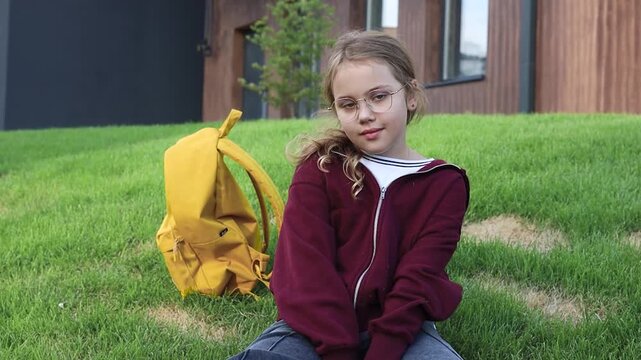 Caucasian child girl sitting at green lawn with yellow backpack smiling to camera.Schoolgirl.Bacl to school. Pupil.