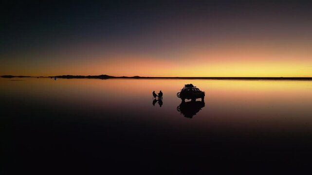 Drone footage circling 4x4 vehicle and two people on chairs at salt flats during sunset. Perfect mirror reflection on water surface. Mountains silhouetted on horizon at dusk.