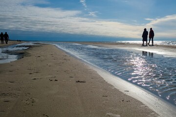 People walking along a sandy beach near the shoreline on a sunny day with reflections of sunlight on the water.