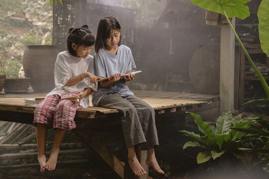 Two Asian children read together on a wooden porch in a rural Thai home, surrounded by warm light, rustic atmosphere, and lush plants. Real Asian countryside lifestyle.