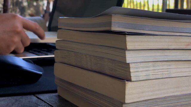a person reading a book in front of a laptop with several stacks of books.