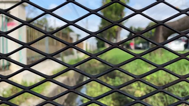 Black metal fence with diamond pattern, blurred greenery in background. Closeup view.